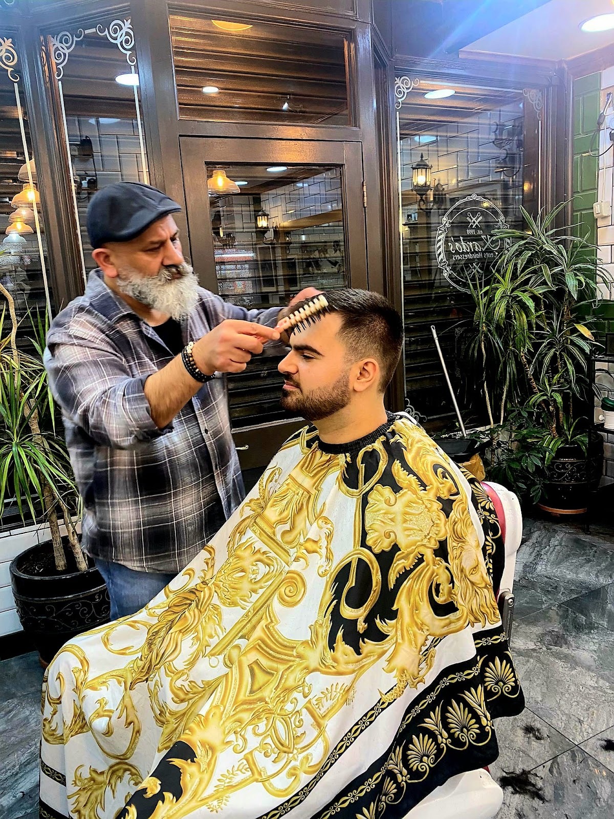 The bearded barber combing a seated client's hair in the plant-filled shop